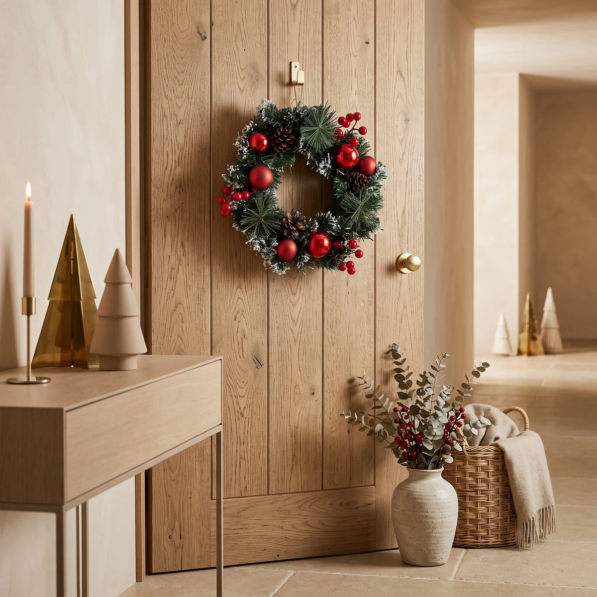 Guirlanda de Natal com bolas vermelhas, pinhas e neve decorando porta de madeira em ambiente rústico sofisticado, com árvores douradas, castiçal e vaso com eucalipto