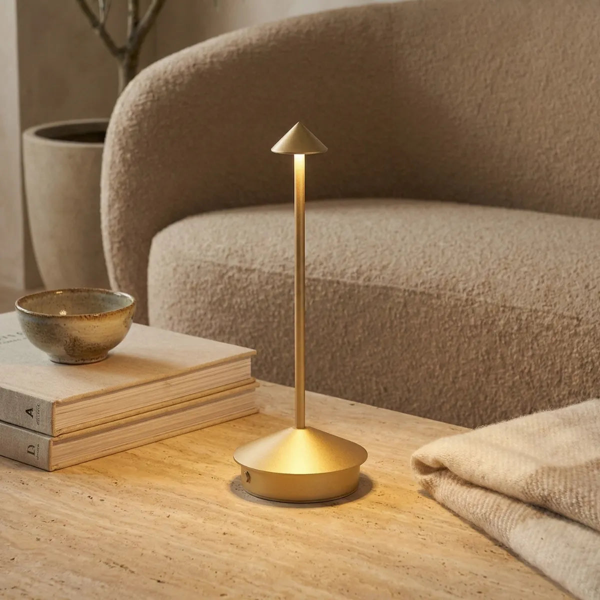 Luminária de mesa sem fio Apollo dourada iluminando mesa de centro de pedra travertino ao lado de sofá curvo moderno.