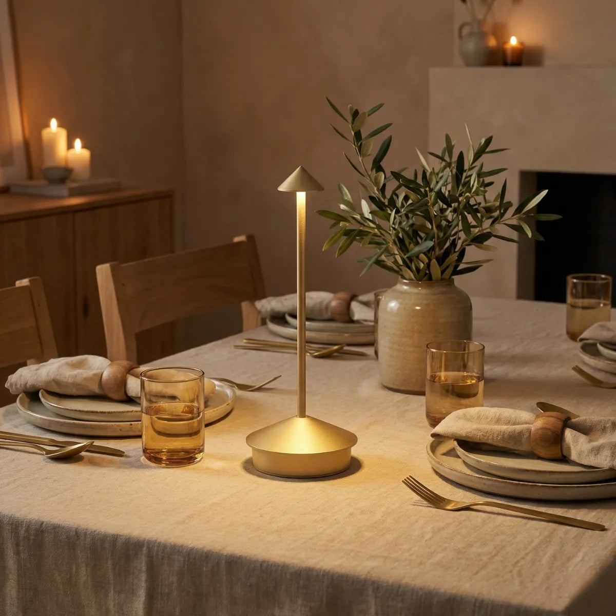 Luminária de mesa posta Apollo dourada decorando jantar elegante com toalha de linho, vaso de cerâmica e arranjo de folhas.