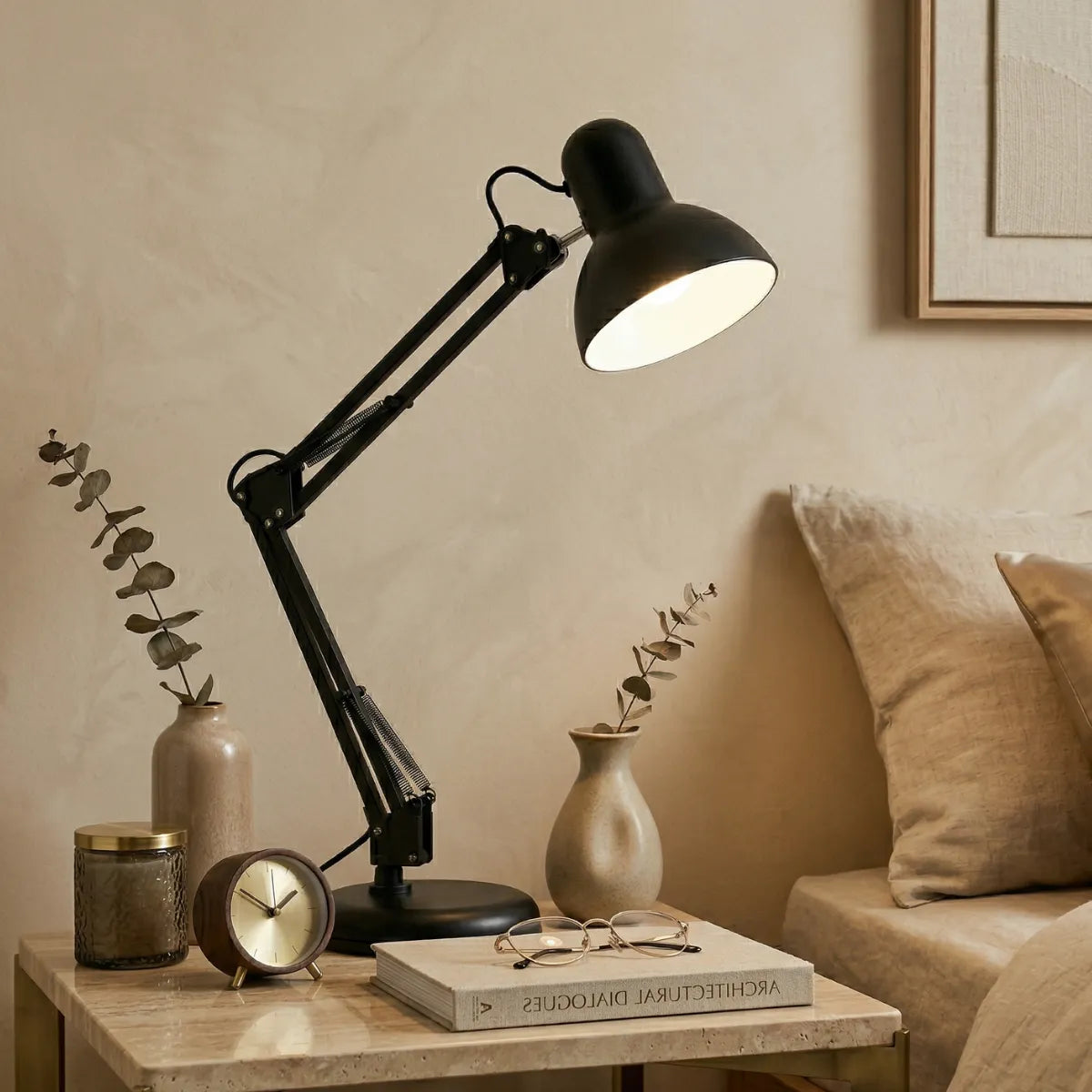 Luminária de mesa Artic preta posicionada sobre uma mesa de cabeceira, ao lado de uma cama com roupa de cama branca e uma planta decorativa ao fundo, criando um ambiente de leitura aconchegante.