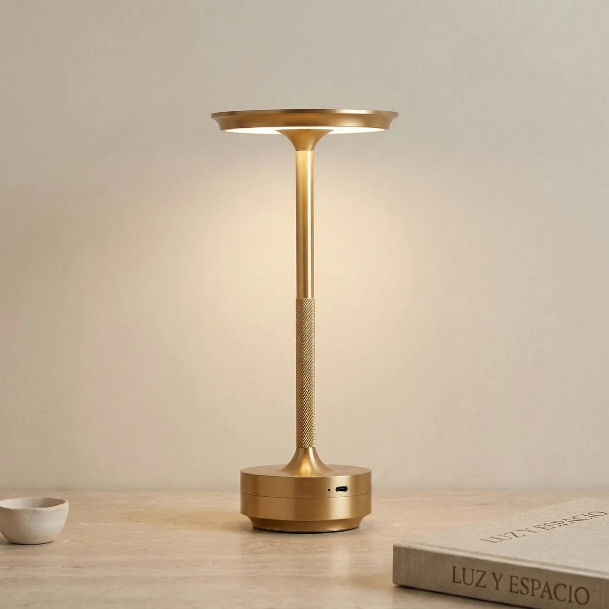 Luminária de mesa Áurea dourada acesa, com design moderno de cúpula plana e haste texturizada, sobre uma superfície clara ao lado de um livro de decoração.