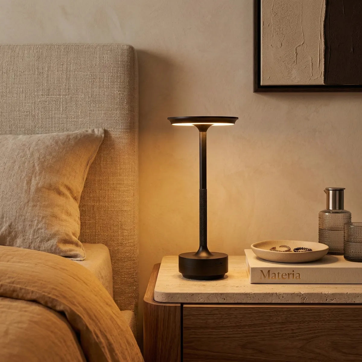 A luminária iluminando uma mesa de cabeceira de madeira ao lado de uma cama. A luz quente destaca um livro e pequenos objetos decorativos, reforçando o uso do produto como luz de leitura ou de apoio no quarto.