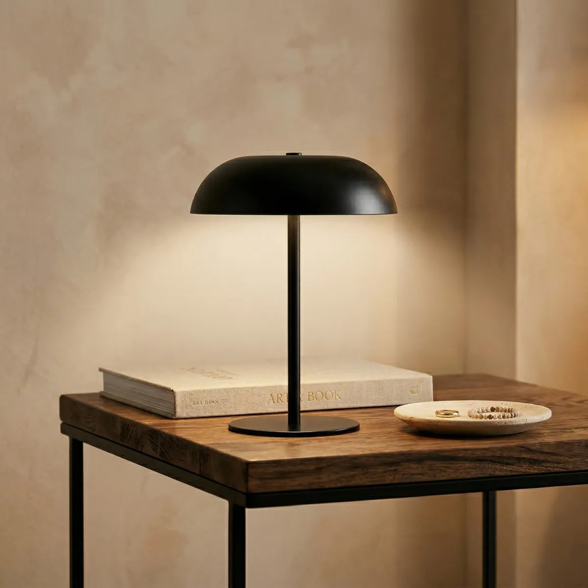 Luminária de mesa Doma preta acesa sobre uma mesa lateral que combina tampo de madeira rústica e estrutura de metal escuro. Ao lado da base, repousam um grande livro de arte e um pequeno prato de pedra clara com joias, contra um fundo de parede bege texturizada.