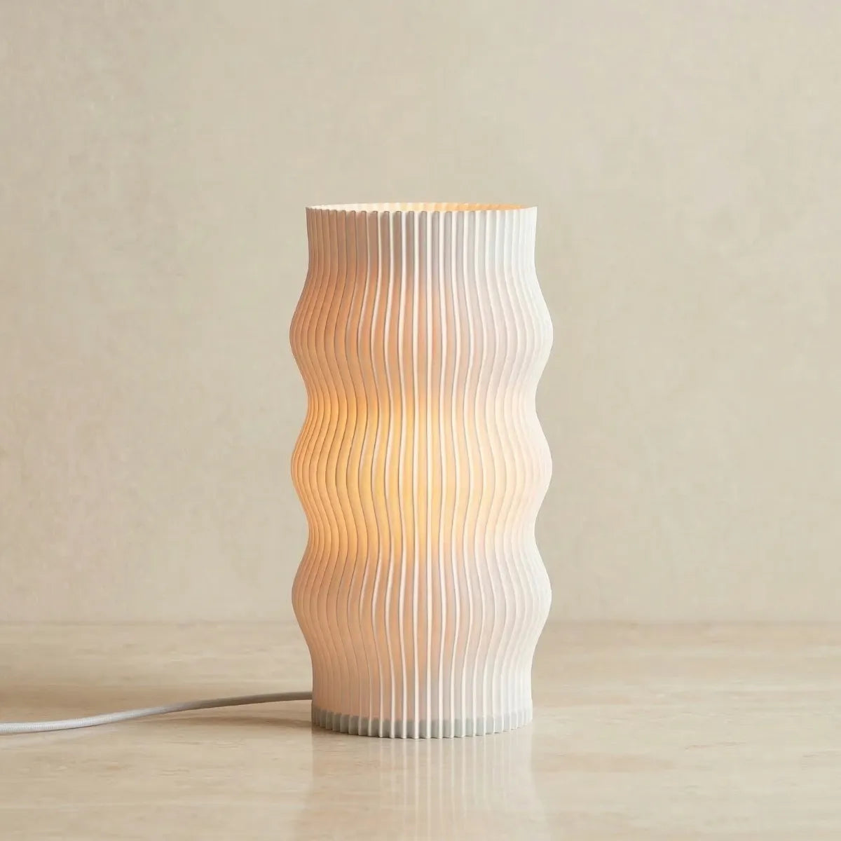 Luminária de mesa Duna acesa, cor branca. A peça possui design cilíndrico com formato orgânico ondulado e textura plissada vertical, posicionada sobre uma superfície lisa e clara.