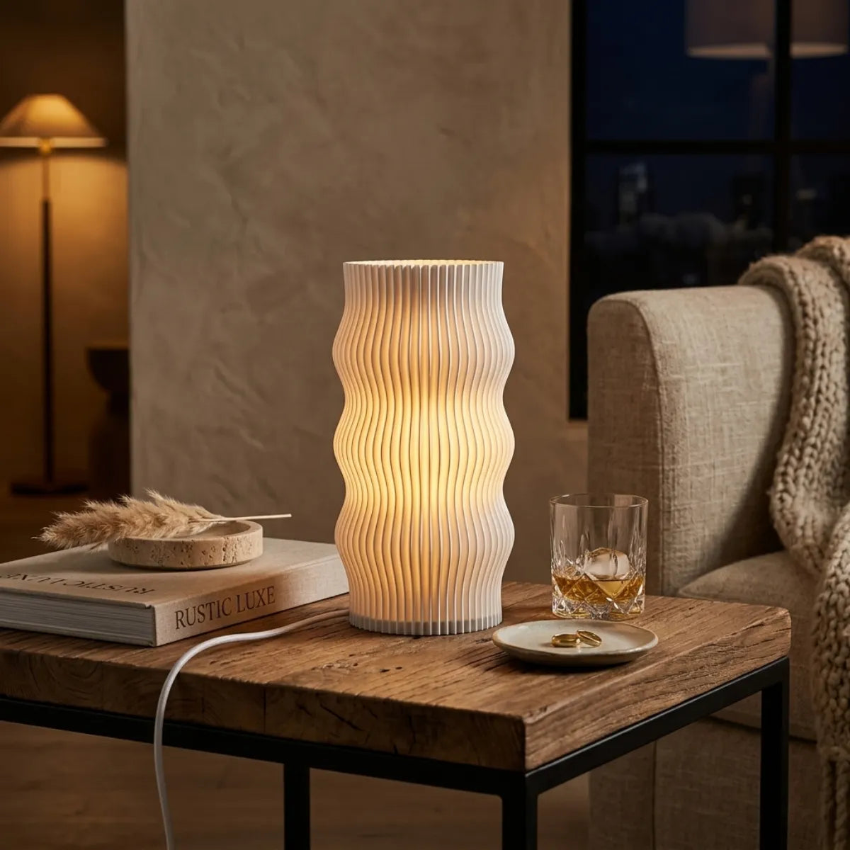 Atmosfera intimista noturna com a luminária Duna sobre uma mesa rústica de madeira e metal. A luz quente destaca um copo de whisky com gelo, pratinhos de cerâmica e o livro "Rustic Luxe".