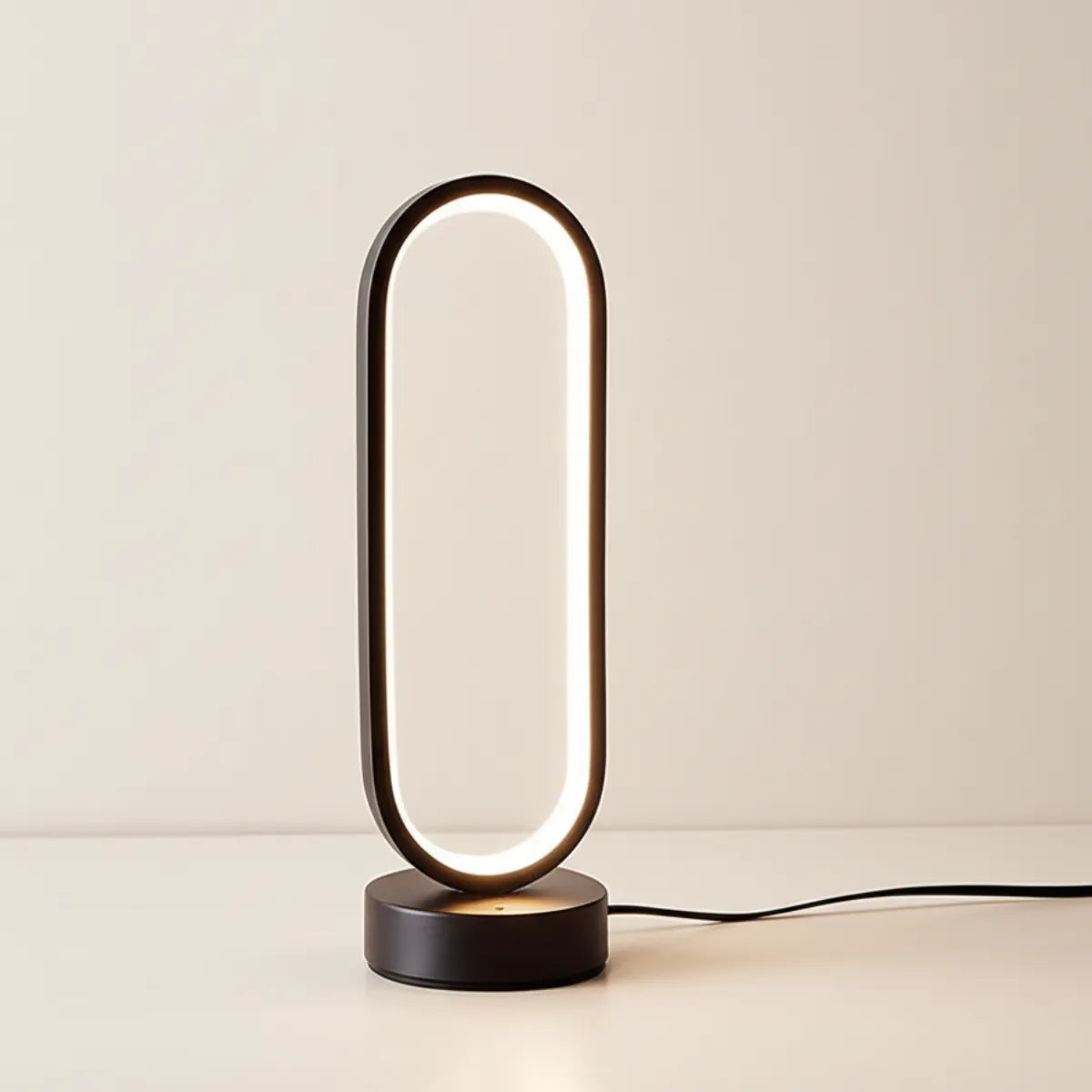 Luminária de mesa Elo preta com design oval minimalista, acesa com luz branca suave e cabo preto, em fundo claro.