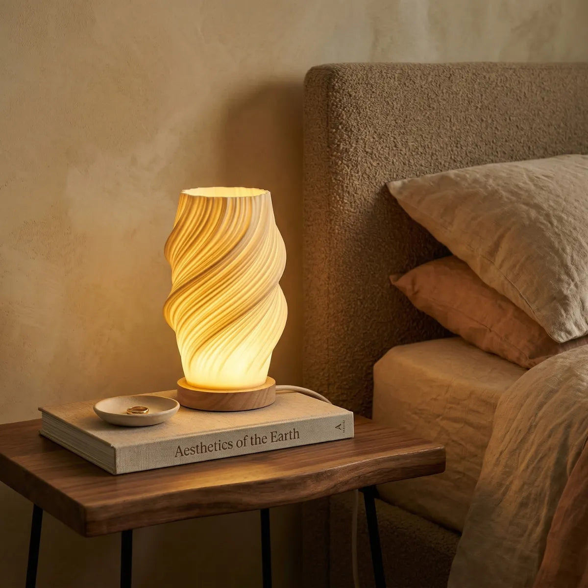 Luminária de mesa acesa com cúpula branca canelada, sobre uma mesa de cabeceira de madeira em um quarto, sobre um livro.