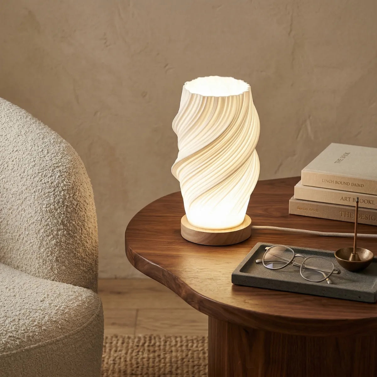Luminária de mesa acesa com cúpula branca canelada, em uma mesa lateral de madeira rústica. Ao lado, três livros, óculos e bandeja. Fundo com poltrona acolhedora.