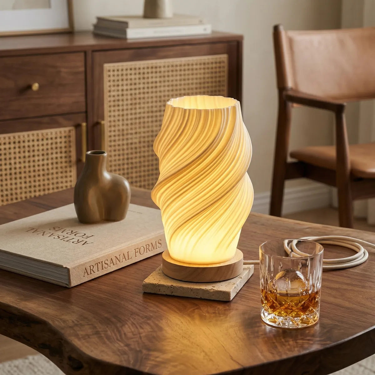 Luminária de mesa Ondea acesa, emitindo luz quente e suave, posicionada sobre uma mesa de centro. A cúpula cônica branca brilha por dentro. Ao lado dela, um livro e um pequeno vaso.