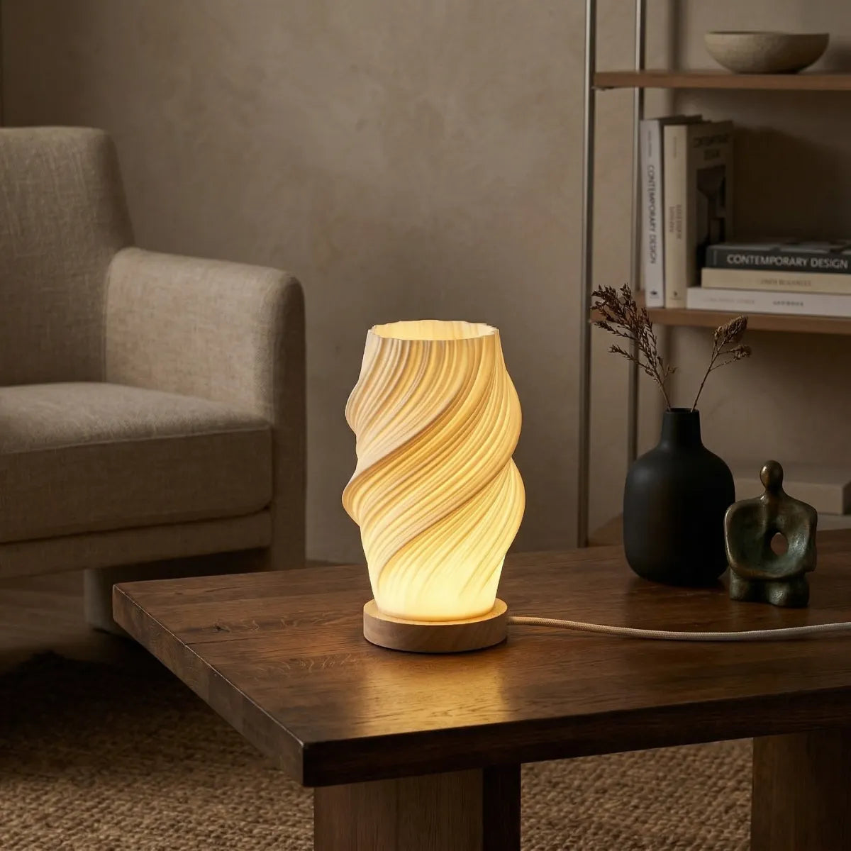 Luminária de mesa Ondea acesa de noite. Apresenta base circular plana de cor natural clara, com cúpula cônica branca. Está sobre uma mesa de centro retangular madeira ao lado de vaso. Poltrona bege ao fundo.