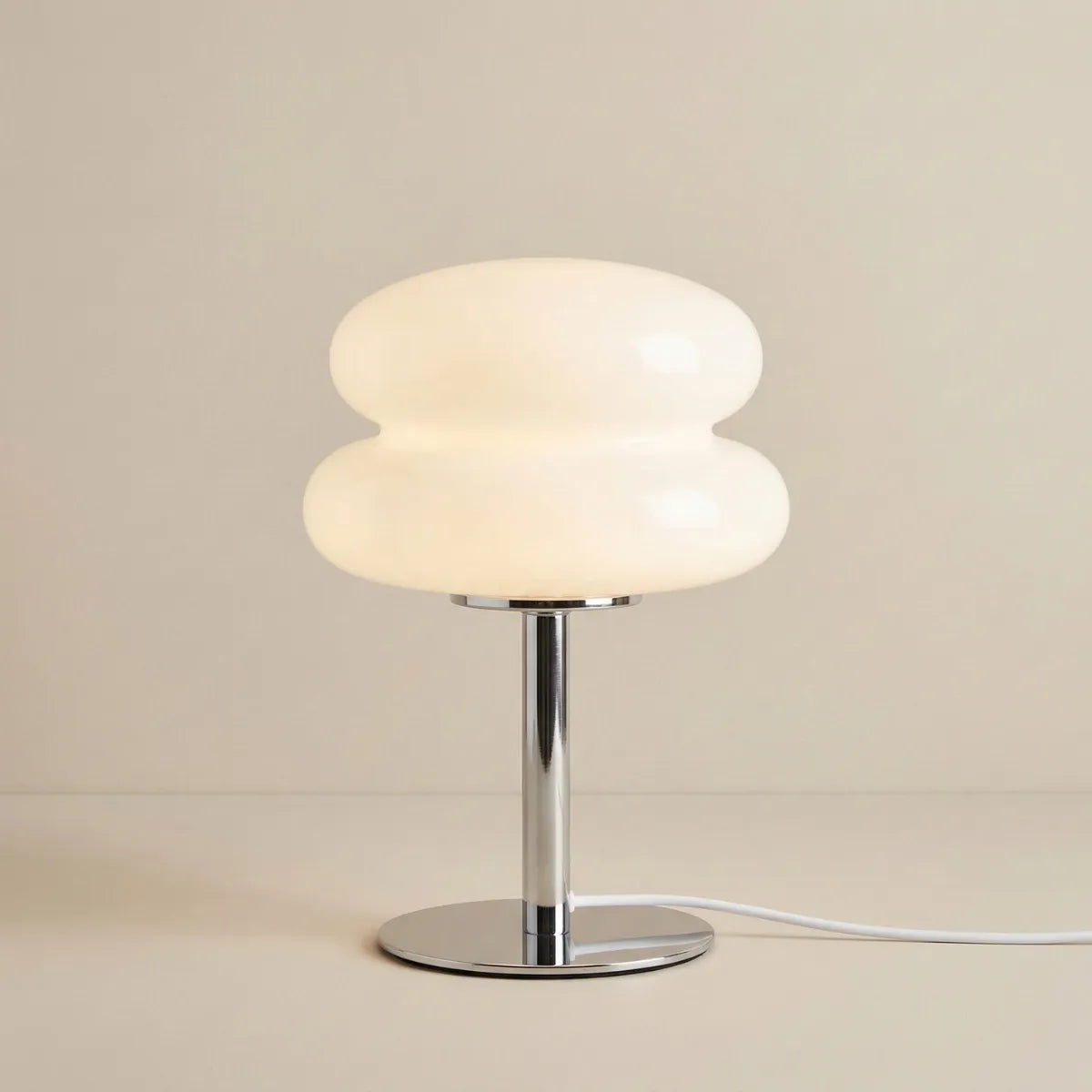 Luminária de mesa Orbe acesa, com cúpula branca em formato orgânico que lembra duas esferas achatadas. A base plana e a haste cilíndrica possuem acabamento em metal cromado, sobre fundo bege minimalista.