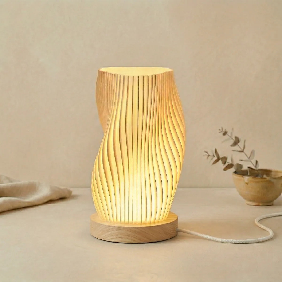 Luminária de mesa Velae acesa, revelando a textura ondulada e a base circular de madeira. Ela está em uma superfície de mesa bege neutra, com um pano de linho e um vaso cerâmico com galhos de eucalipto seco ao lado, contra uma parede texturizada.