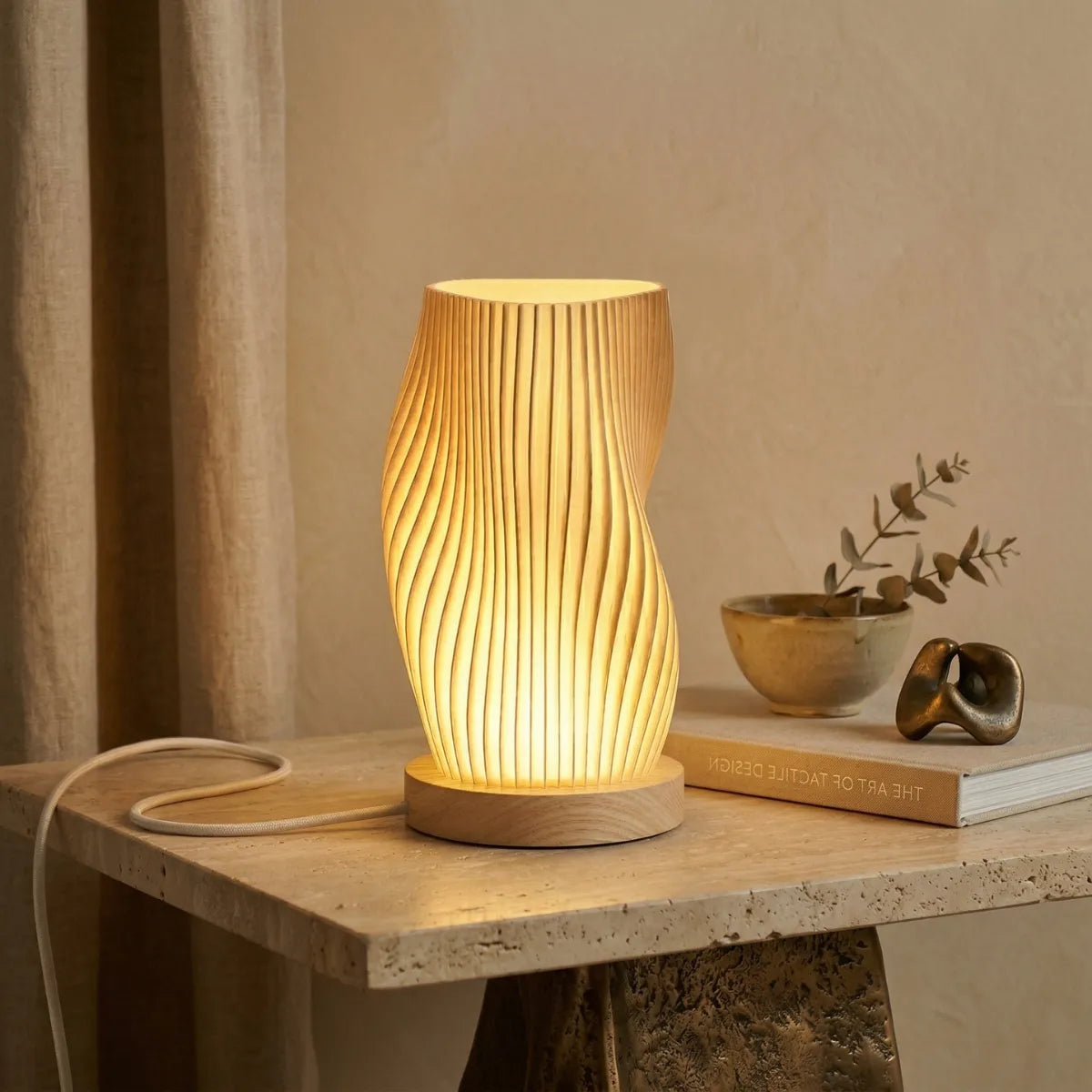 Close-up de uma luminária de mesa Velae acesa em uma mesa lateral de travertino com uma base de metal texturizado. Ao lado dela, há um livro, um pequeno vaso de eucalipto e uma escultura dourada abstrata de nó, com uma cortina de linho visível à esquerda.