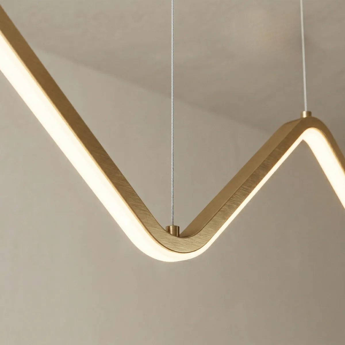 Detalhe em macro da luminária pendente dourada. A imagem destaca o acabamento metálico escovado na parte superior da estrutura em zigue-zague e a faixa contínua de luz de LED branca acesa na parte inferior.