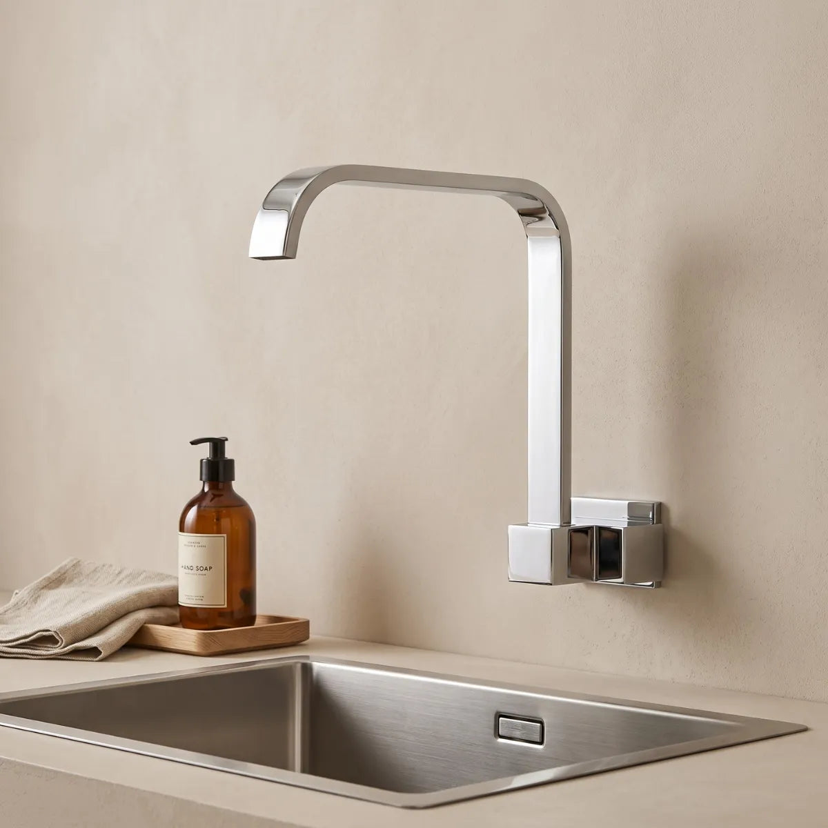Torneira para cozinha de parede cromada Cubus com bica quadrada em ângulo reto e acabamento espelhado, instalada sobre cuba de inox com saboneteira âmbar e bandeja de madeira.