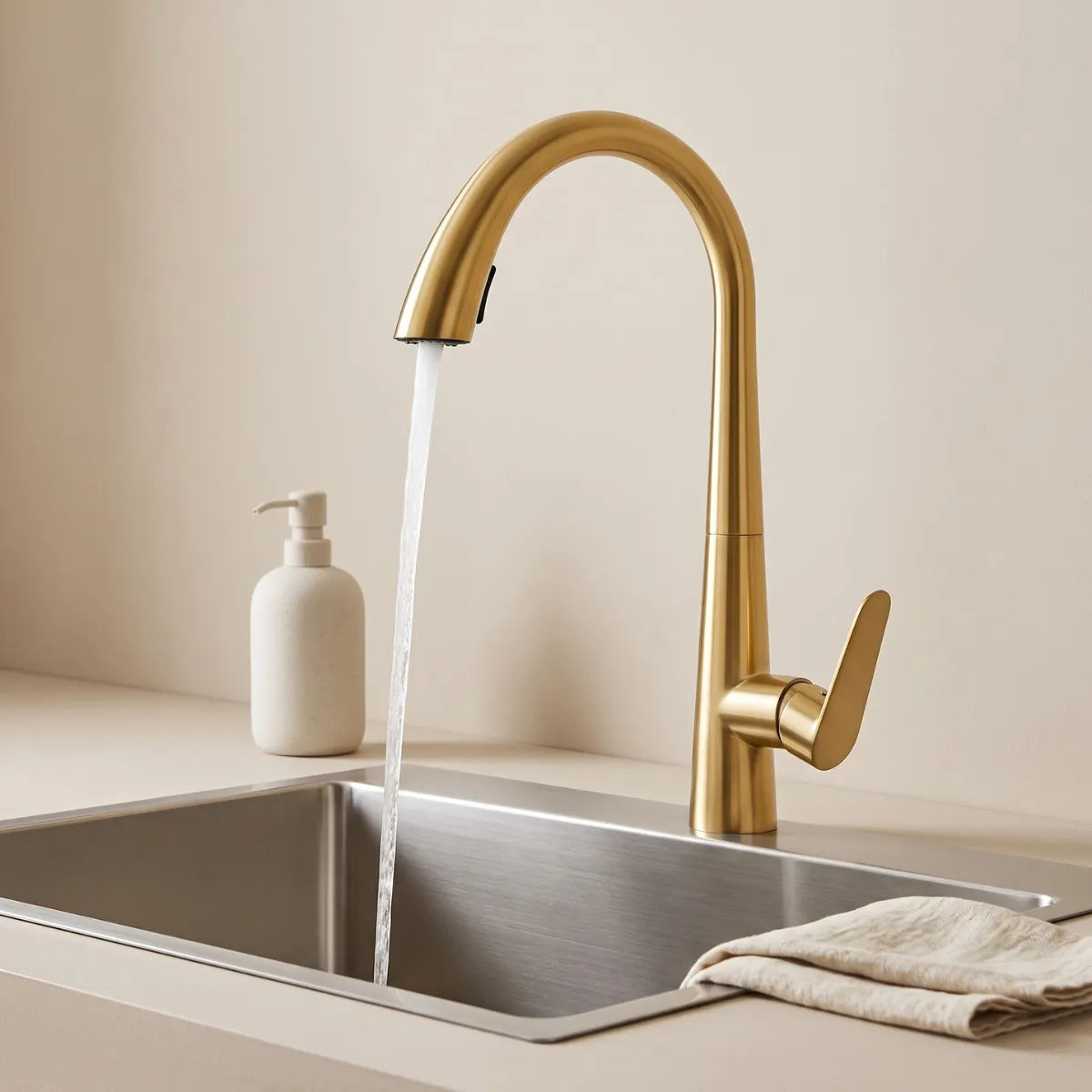 Torneira para cozinha gourmet dourada Cygne com bica em arco orgânico e jato concentrado ativo sobre cuba de inox, bancada neutra com saboneteira e pano de linho.