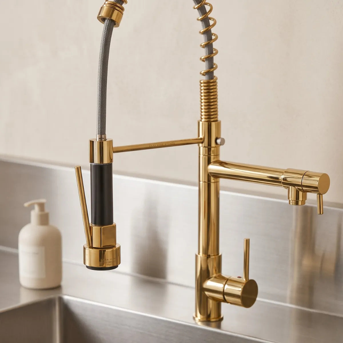 Detalhe da ducha e da bica articulada da torneira para cozinha dourada Bocuse, com acabamento bicolor ouro e preto e mola espiral texturizada sobre cuba de inox.