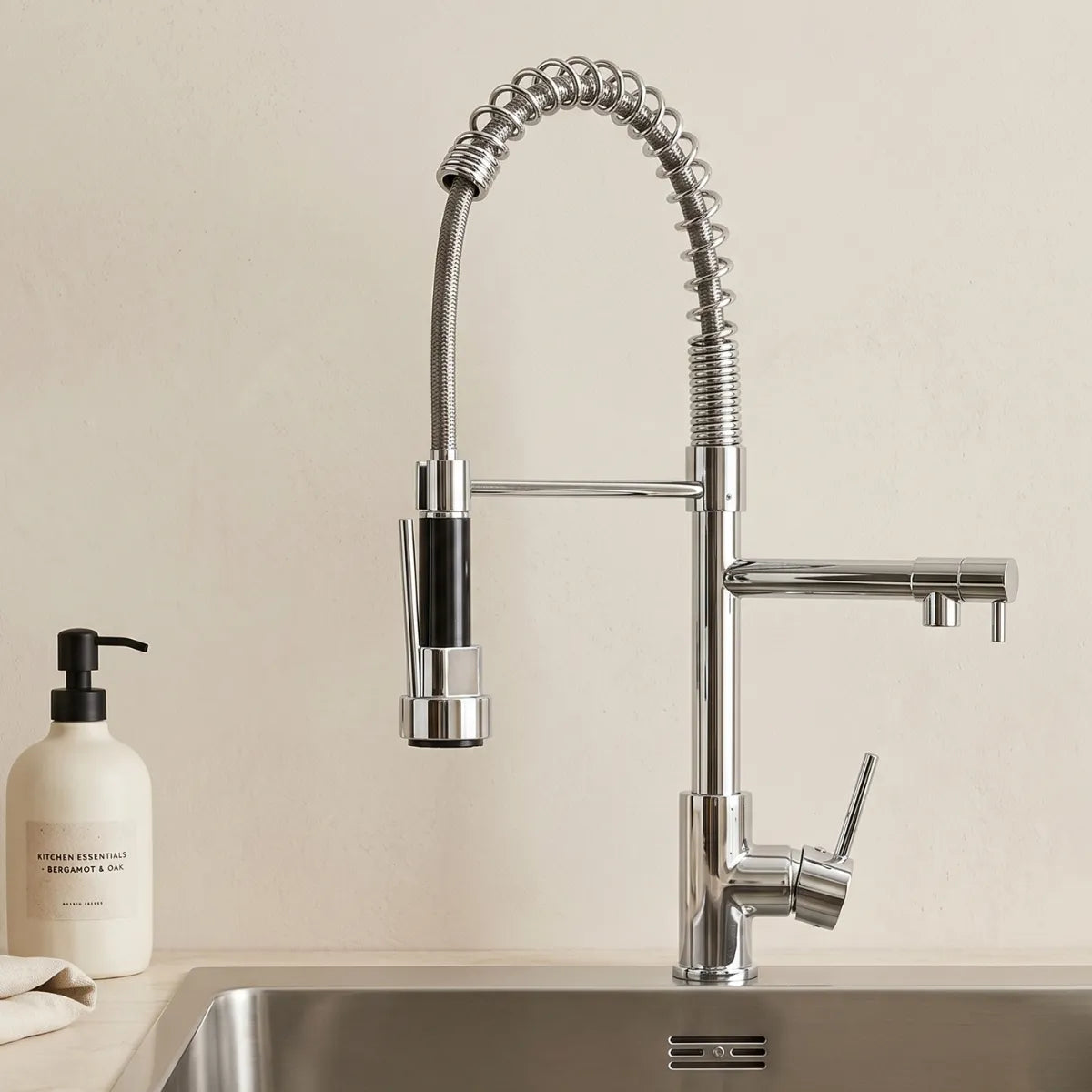 Torneira para cozinha gourmet cromada monocomando com mola espiral e ducha flexível recolhida, instalada sobre cuba de inox com saboneteira bege ao lado, fundo neutro.
