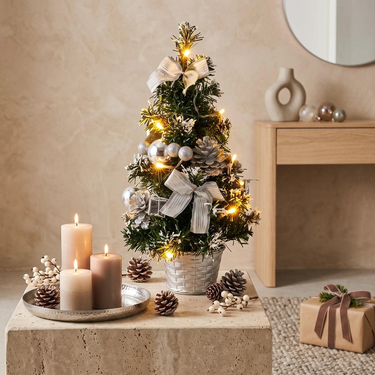 Árvore de Natal pequena iluminada com laços prateados e pinhas decorando mesa de mármore em ambiente rústico sofisticado, com velas, presentes embrulhados e vaso de cerâmica