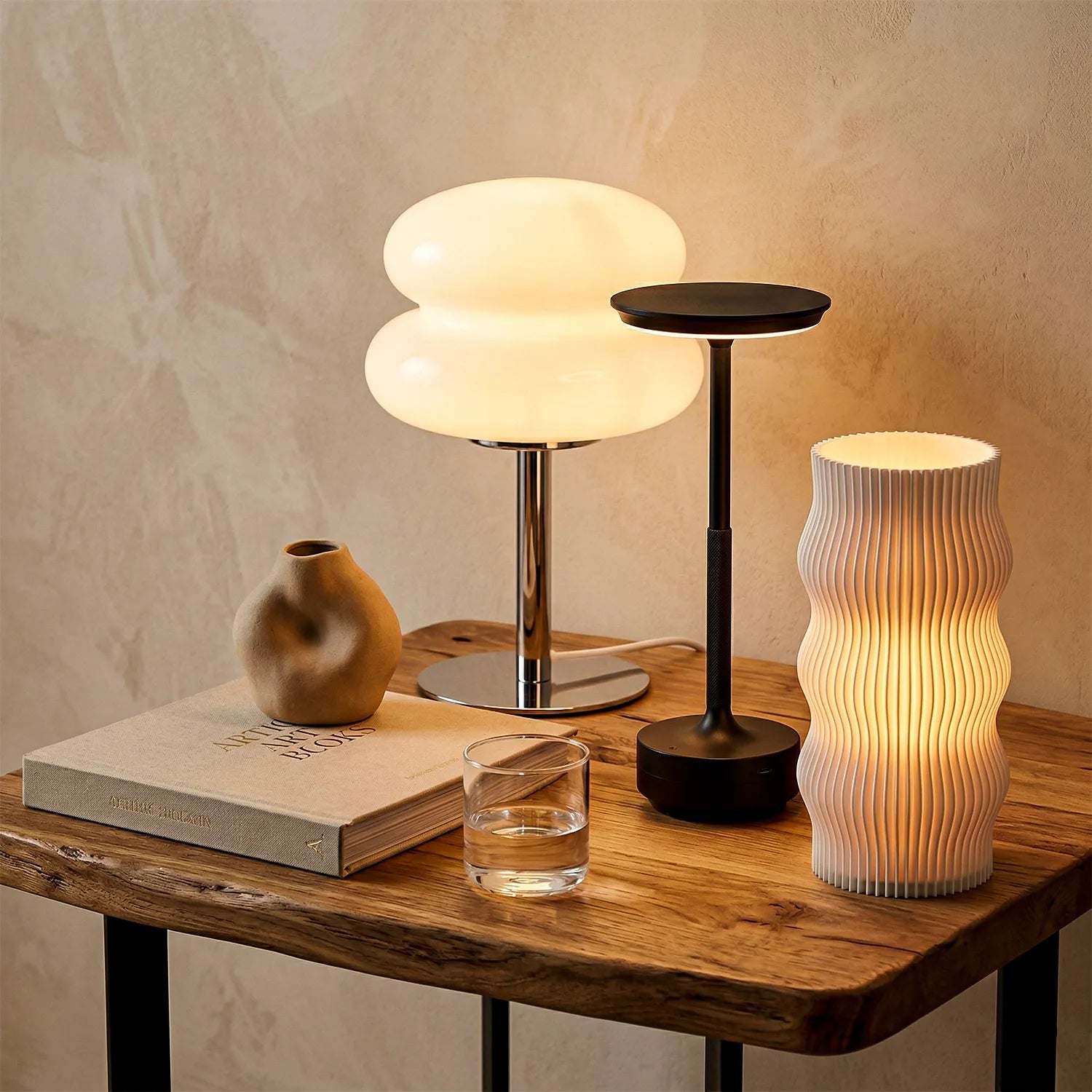 Mesa lateral rústica com 3 luminárias de mesa, acesas. Acompanha livro, vaso orgânico e copo de água contra parede clara texturizada.