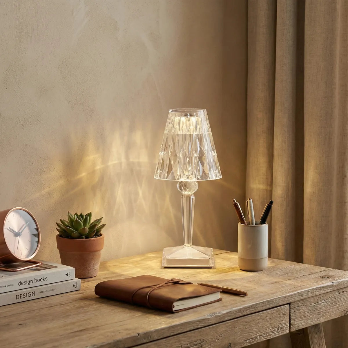 Abajur de mesa em acrílico cristal iluminando escrivaninha de madeira com cadernos e plantas, adicionando um toque de glamour ao home office.