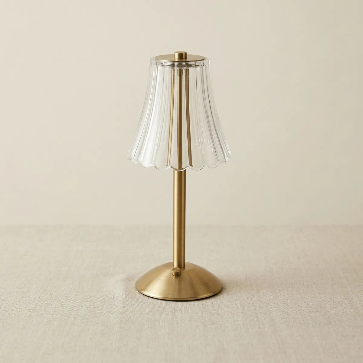 Abajur de mesa dourado desligado, com base metálica e cúpula transparente canelada em formato de sino, sobre fundo bege liso.