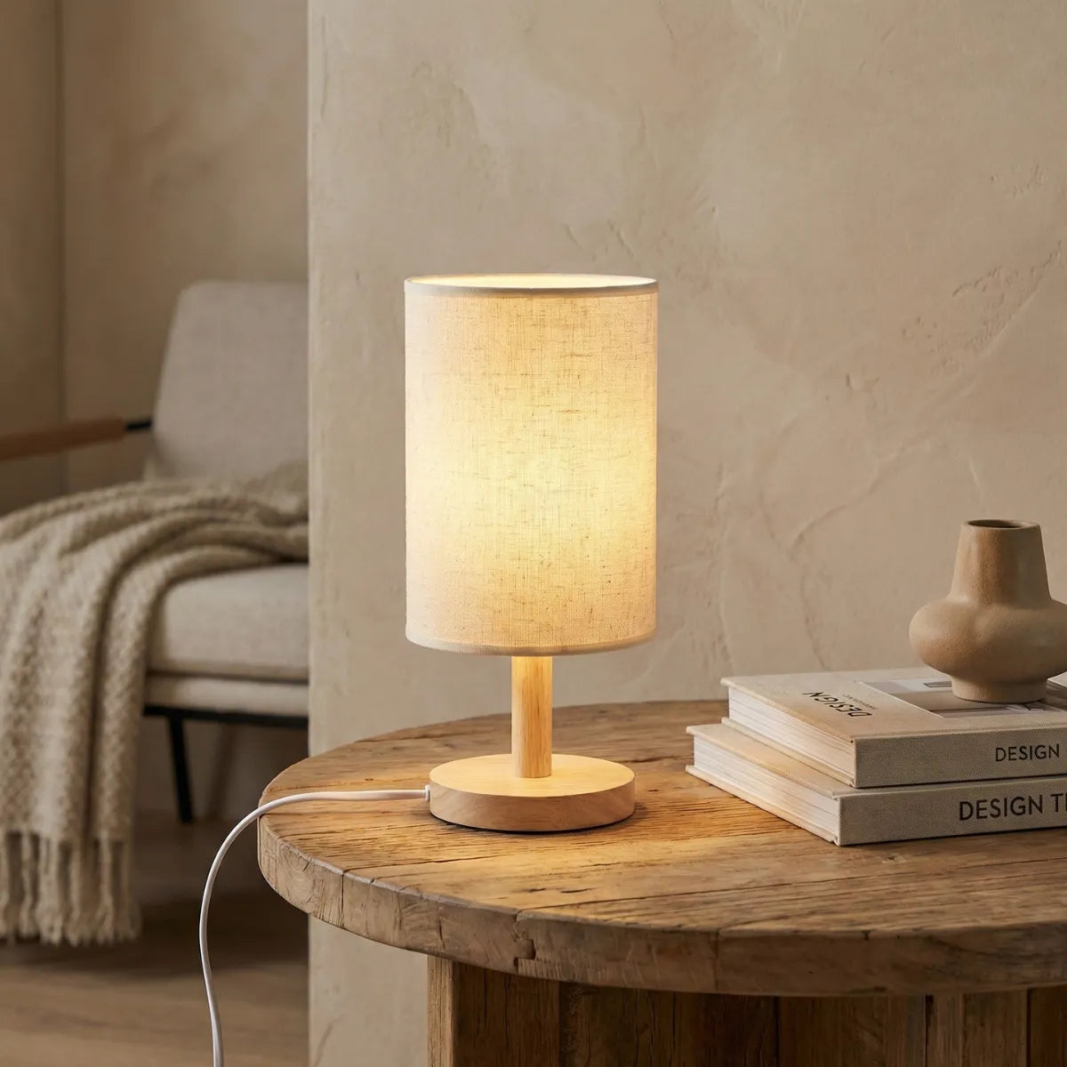 Abajur de mesa com base de madeira Fuste iluminando canto de leitura com livros de design e poltrona confortável.