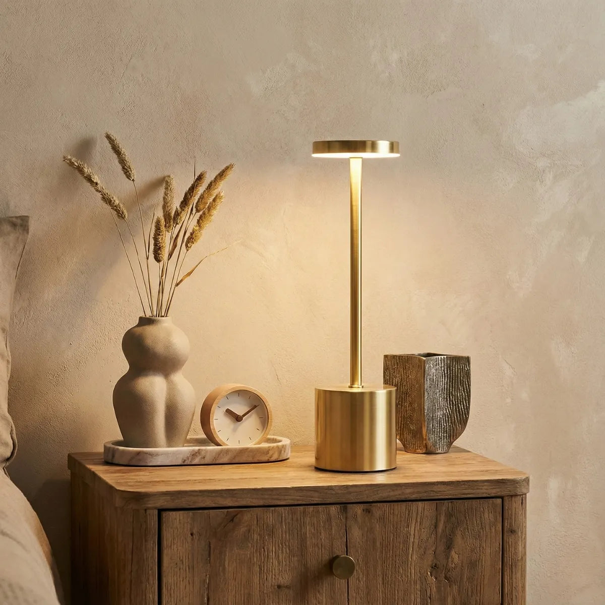 Luminária de mesa dourada na mesa de cabeceira de madeira, criando iluminação acolhedora em quarto de casal com tons terrosos.