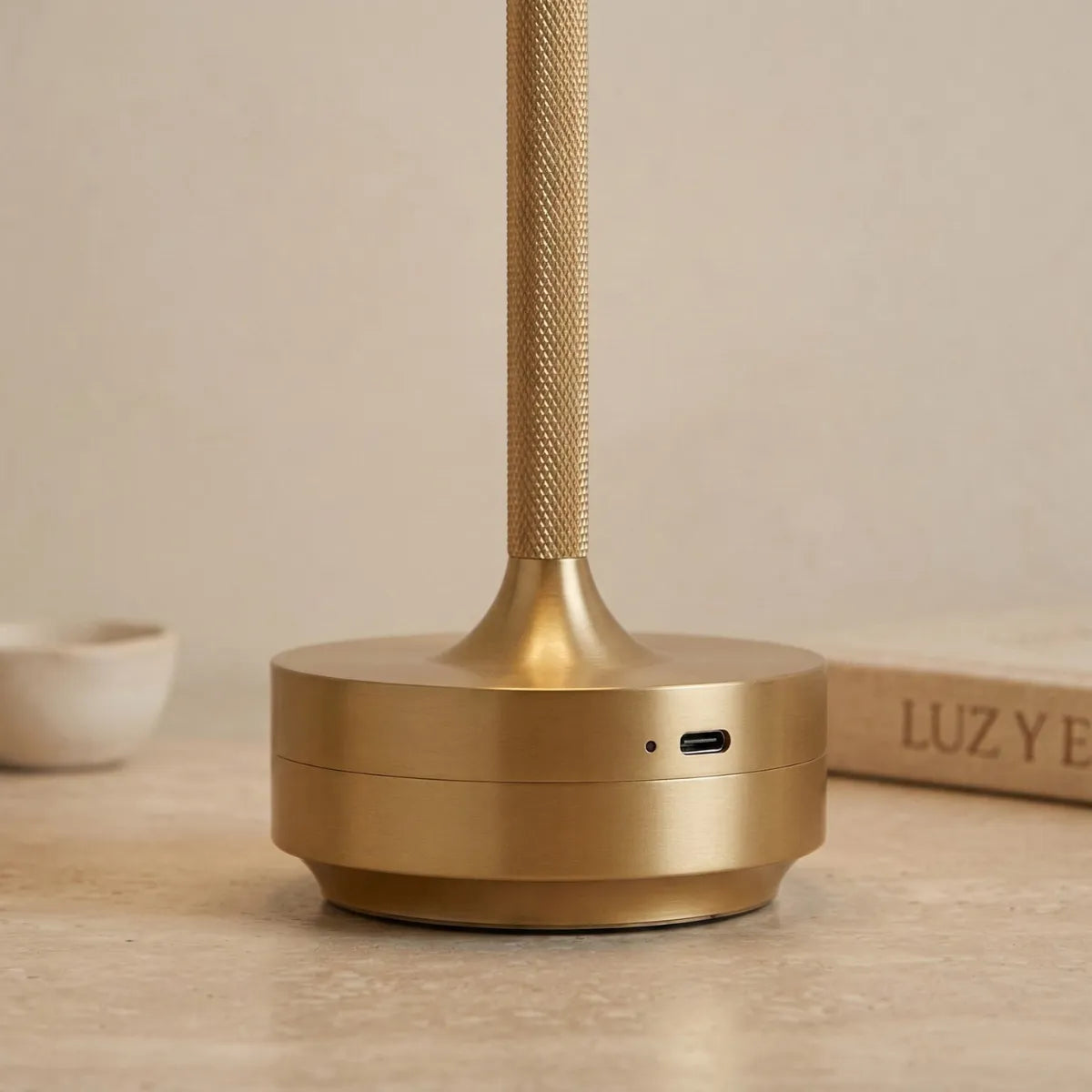 Detalhe em close da base circular da luminária Áurea dourada, destacando o acabamento metálico fosco elegante e a entrada USB-C para carregamento.