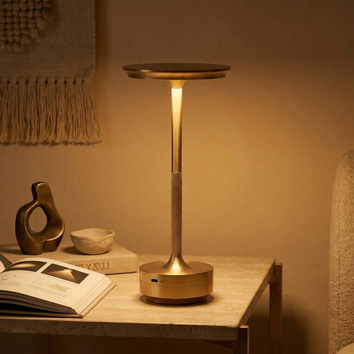 Luminária de mesa Áurea dourada acesa ao lado de um livro aberto e uma escultura orgânica, destacando sua luz suave e design sofisticado contra uma parede texturizada.