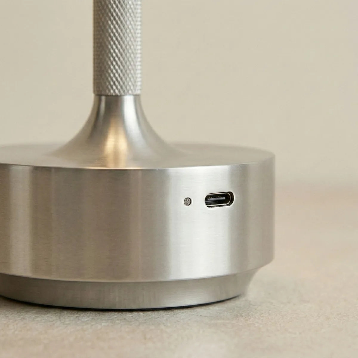 Detalhe do acabamento em metal prata escovado da base da luminária Áurea, destacando a luz de indicação, a entrada USB-C para carregamento e a textura recartilhada da haste.