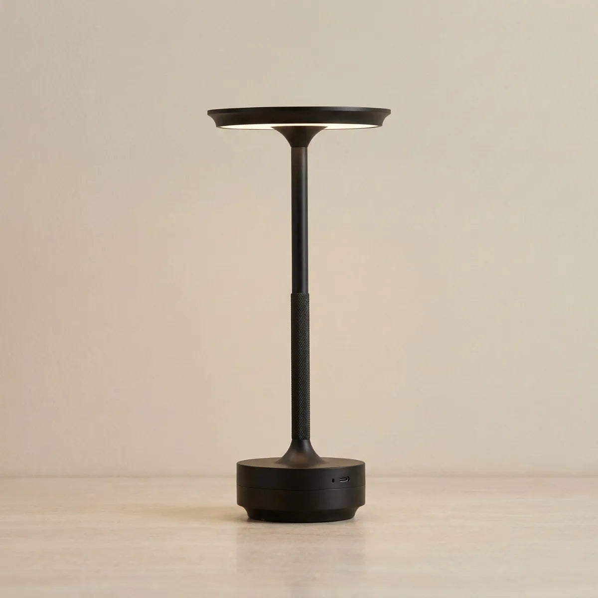 Luminária de mesa Áurea na cor preta com design minimalista e moderno. Possui uma haste fina com detalhe de textura recartilhada no centro, topo circular plano que emite luz para baixo e base cilíndrica robusta. Fundo bege neutro.