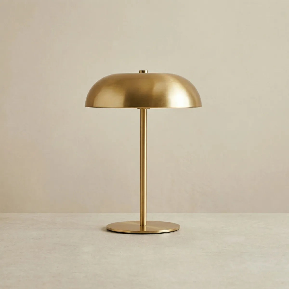 Luminária de mesa Doma dourada apagada e centralizada contra um fundo bege neutro. A foto destaca seu design minimalista em metal escovado, com base circular plana, haste fina e reta, e cúpula lisa em formato de domo.