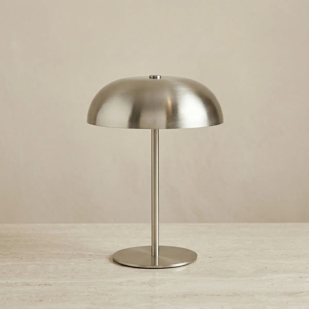 Luminária de mesa Doma prata apagada e centralizada contra um fundo bege neutro. A foto destaca seu design minimalista em metal escovado prateado, com base circular plana, haste fina e reta, e cúpula lisa em formato de domo, apoiada sobre uma superfície de pedra clara.