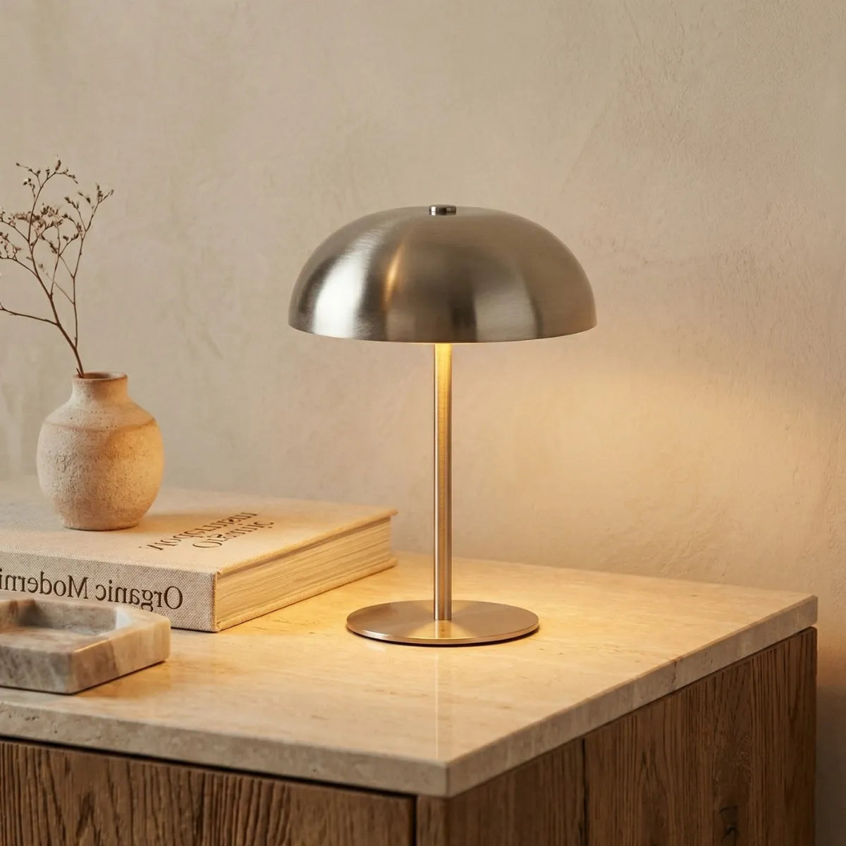 Luminária de mesa Doma prata acesa, criando um clima acolhedor sobre uma bancada de pedra clara e madeira. À esquerda, há um livro servindo de base para um vasinho rústico com uma pequena flor seca, além de uma pequena bandeja de pedra cravada.