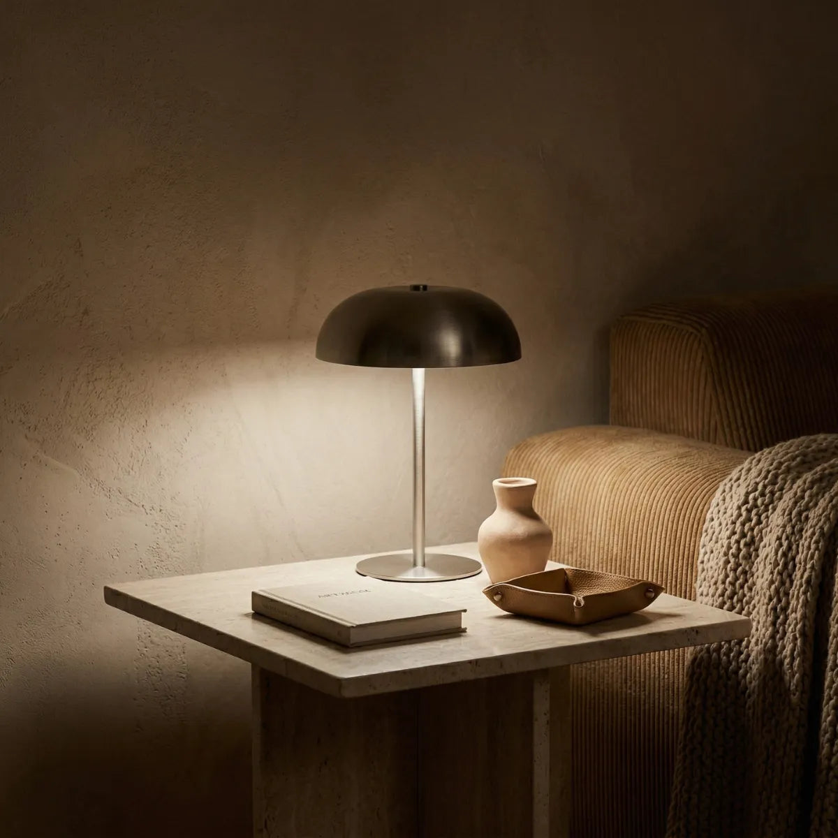 Luminária de mesa Doma prata acesa sobre uma pequena mesa lateral de pedra (travertino). A luz neutra destaca as imperfeições da parede texturizada ao fundo. Acompanham a luminária um livro claro, um pequeno vaso arredondado e uma bandeja organizadora de couro. 