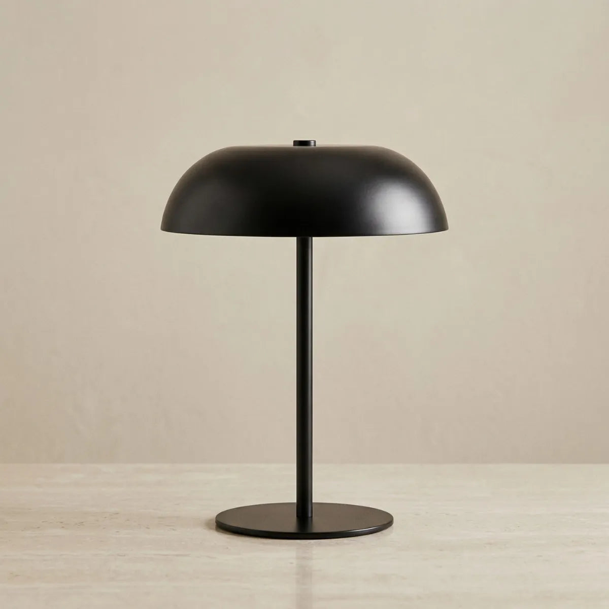 Luminária de mesa Doma preta apagada e centralizada contra um fundo bege neutro. A foto destaca seu design minimalista em metal com acabamento fosco, apresentando uma base circular plana, haste fina e reta, e cúpula lisa em formato de domo.