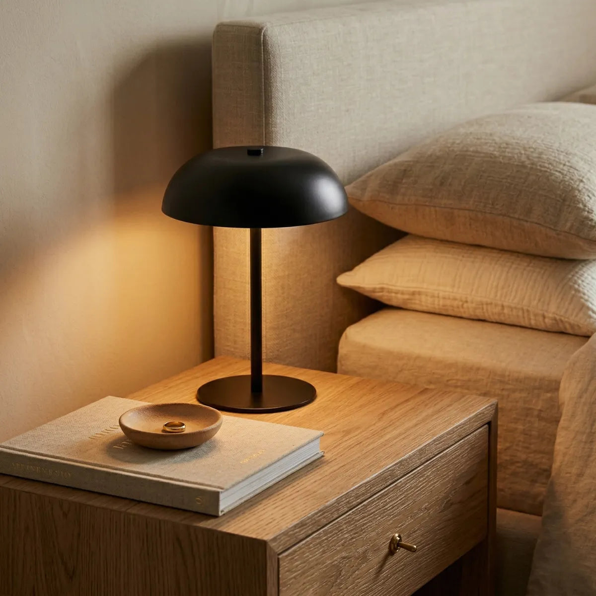 Luminária de mesa Doma preta acesa sobre uma mesa de cabeceira de madeira. A luz quente e aconchegante ilumina um livro de capa dura e um pequeno pratinho de madeira com um anel. À direita, vê-se parte de uma cama arrumada com travesseiros e lençóis de linho claro.