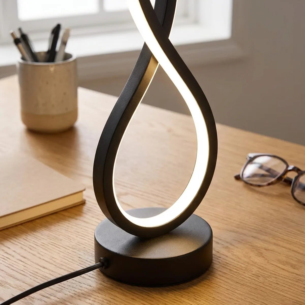 Close-up da Luminária Infinity preta em uma mesa de escritório de madeira, ao lado de óculos, caderno e porta-canetas.