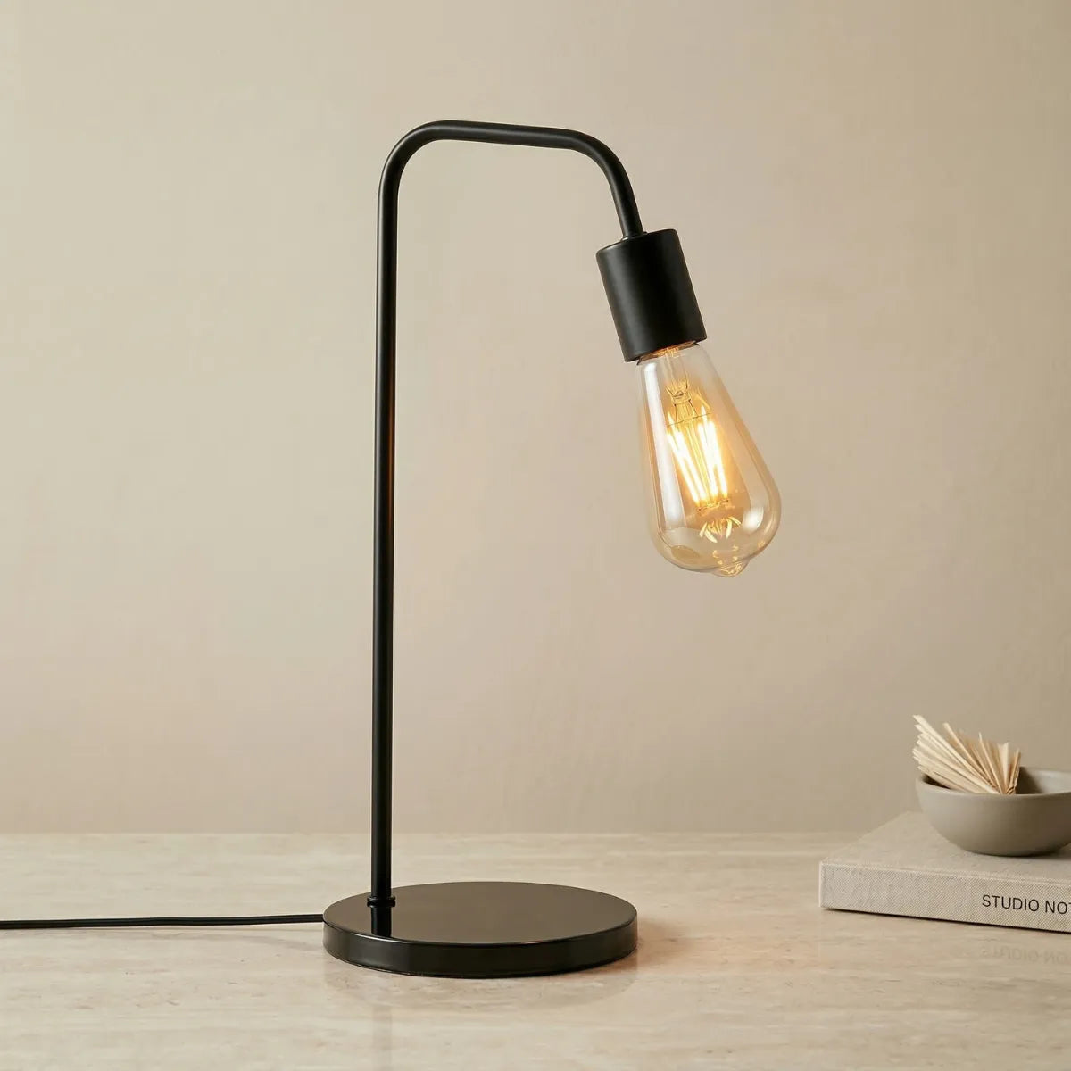 Luminária de mesa Linea preta acesa, com design industrial minimalista. Estrutura tubular em metal fosco curvado, base circular e lâmpada de filamento aparente estilo vintage, sobre uma superfície clara de pedra.
