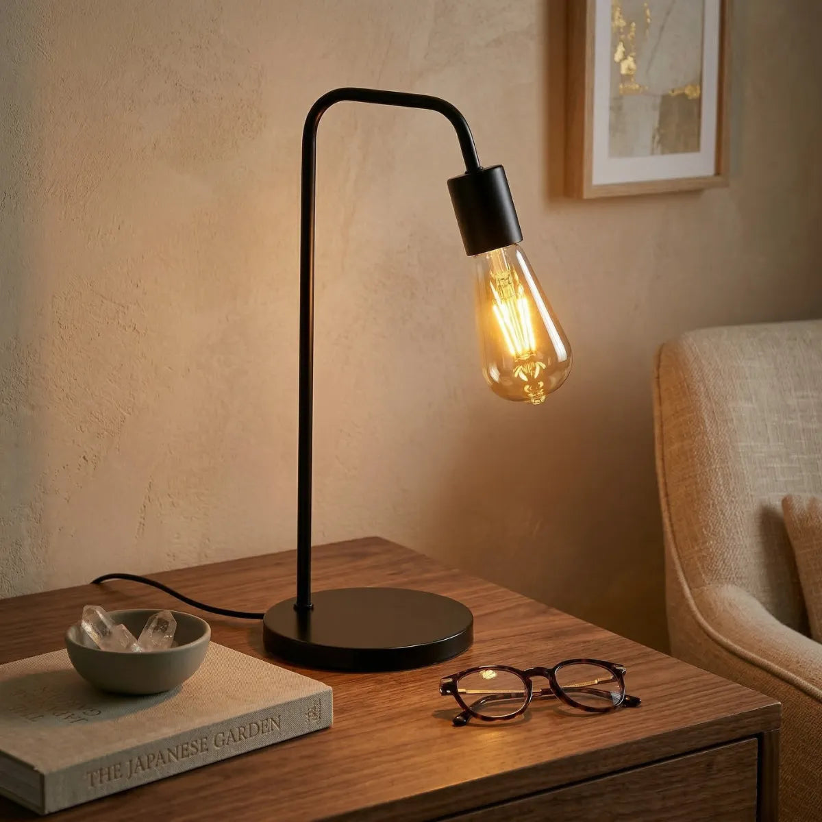 Luminária de mesa Linea preta iluminando suavemente uma mesa de madeira rústica. O design minimalista contrasta com um livro sobre jardins japoneses, óculos de leitura e cristais, criando um canto de leitura intimista.