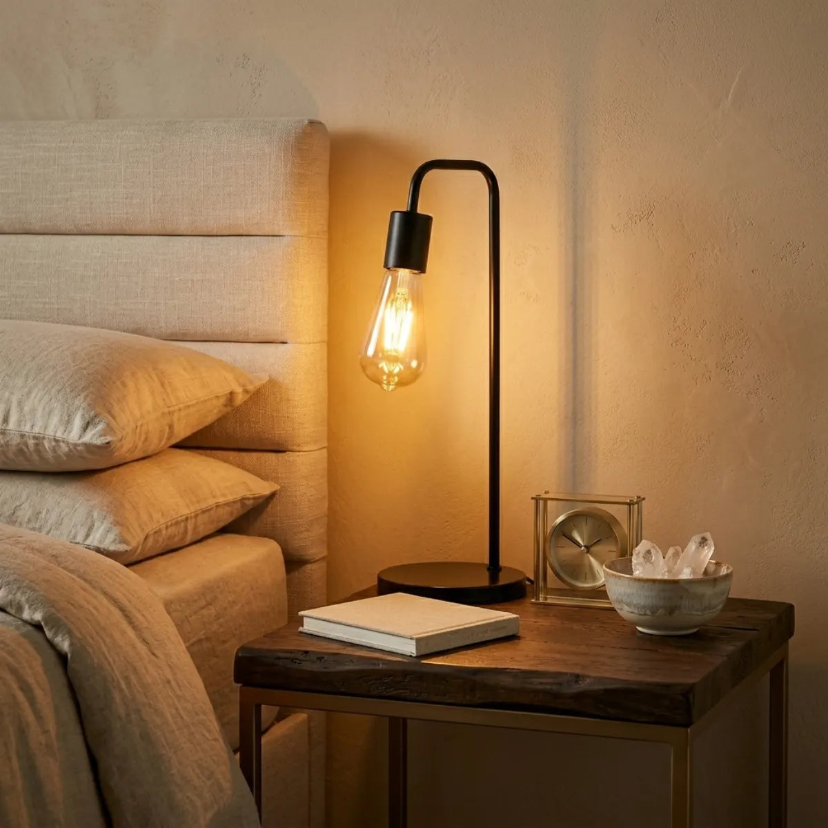 Luminária Linea preta sendo usada como abajur sobre uma mesa de cabeceira de madeira rústica. A luz quente e âmbar da lâmpada de filamento ilumina os travesseiros de uma cama aconchegante e um relógio dourado clássico.