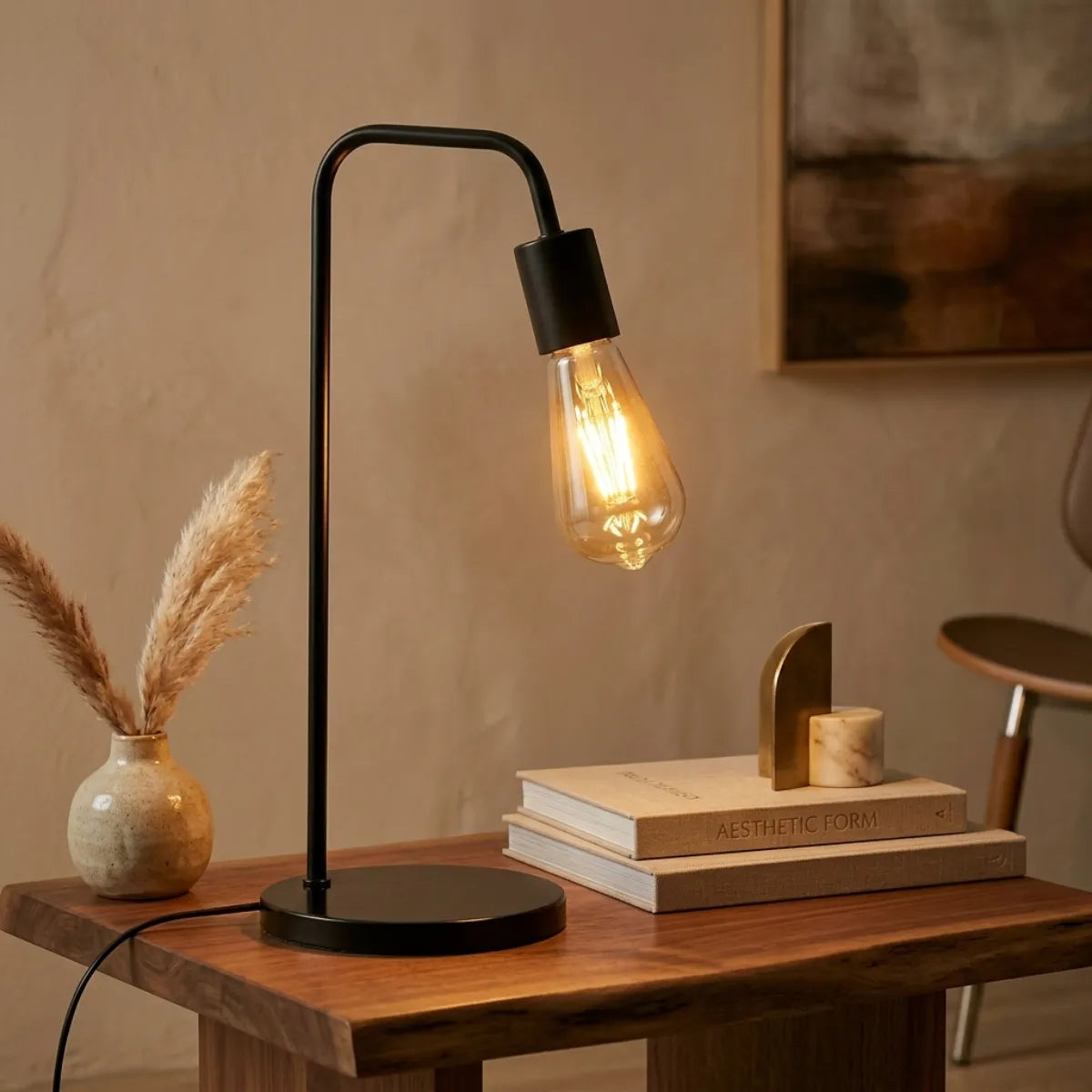 Luminária Linea preta acesa sobre uma mesa lateral de madeira com borda orgânica. A peça ilumina um arranjo de livros de arte, objetos decorativos dourados e um vaso rústico com capim dos pampas, em um ambiente sofisticado em tons neutros.