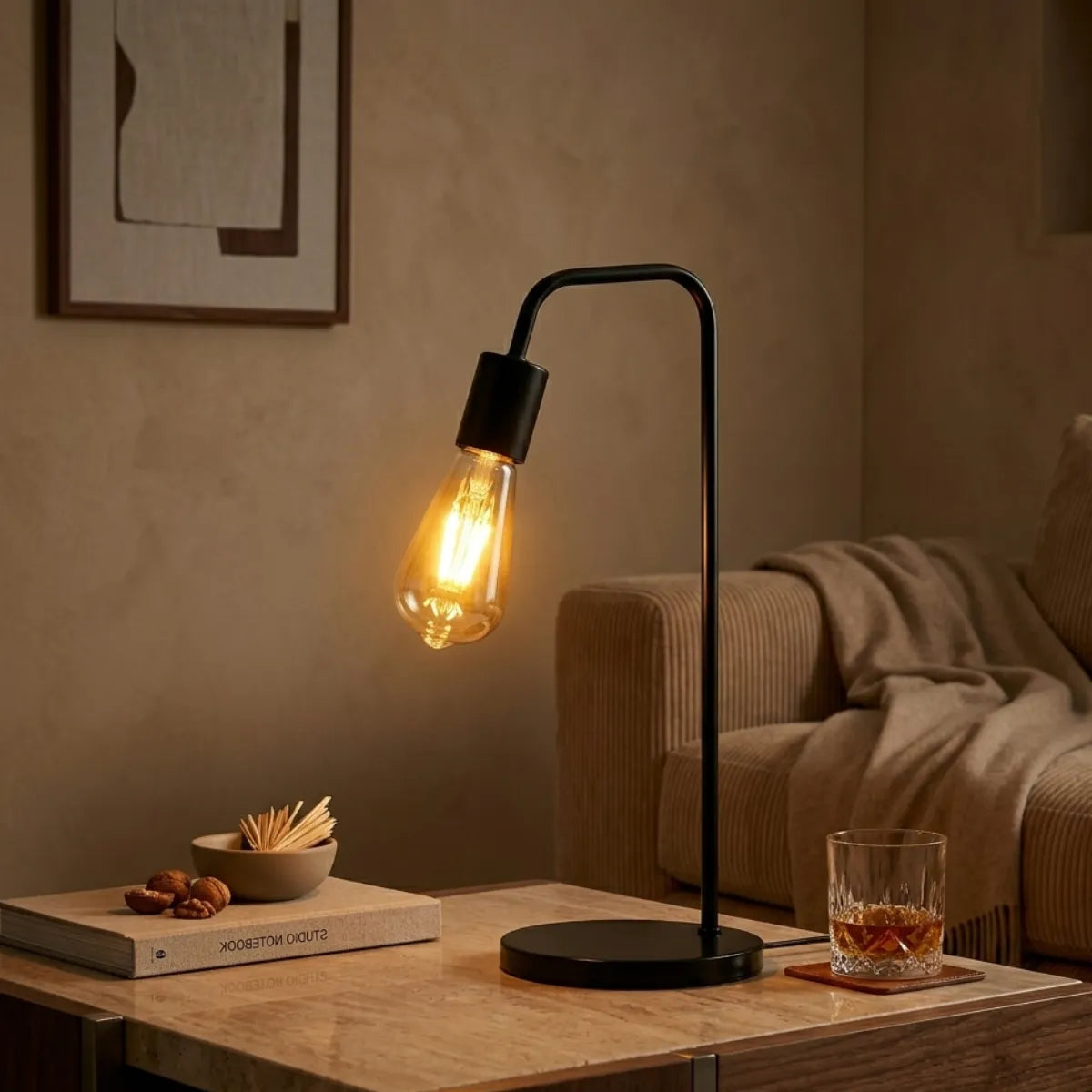 Luminária de mesa Linea com lâmpada retrô iluminando uma mesa de centro de mármore em um clima noturno relaxante. Ao lado da base preta fosca, um livro, pequenos petiscos e um copo de whisky, com um sofá de veludo cotelê ao fundo.