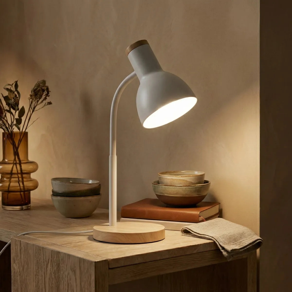 Luminária de mesa Locus branca acesa em prateleira de madeira iluminando livros e decoração em ambiente acolhedor.