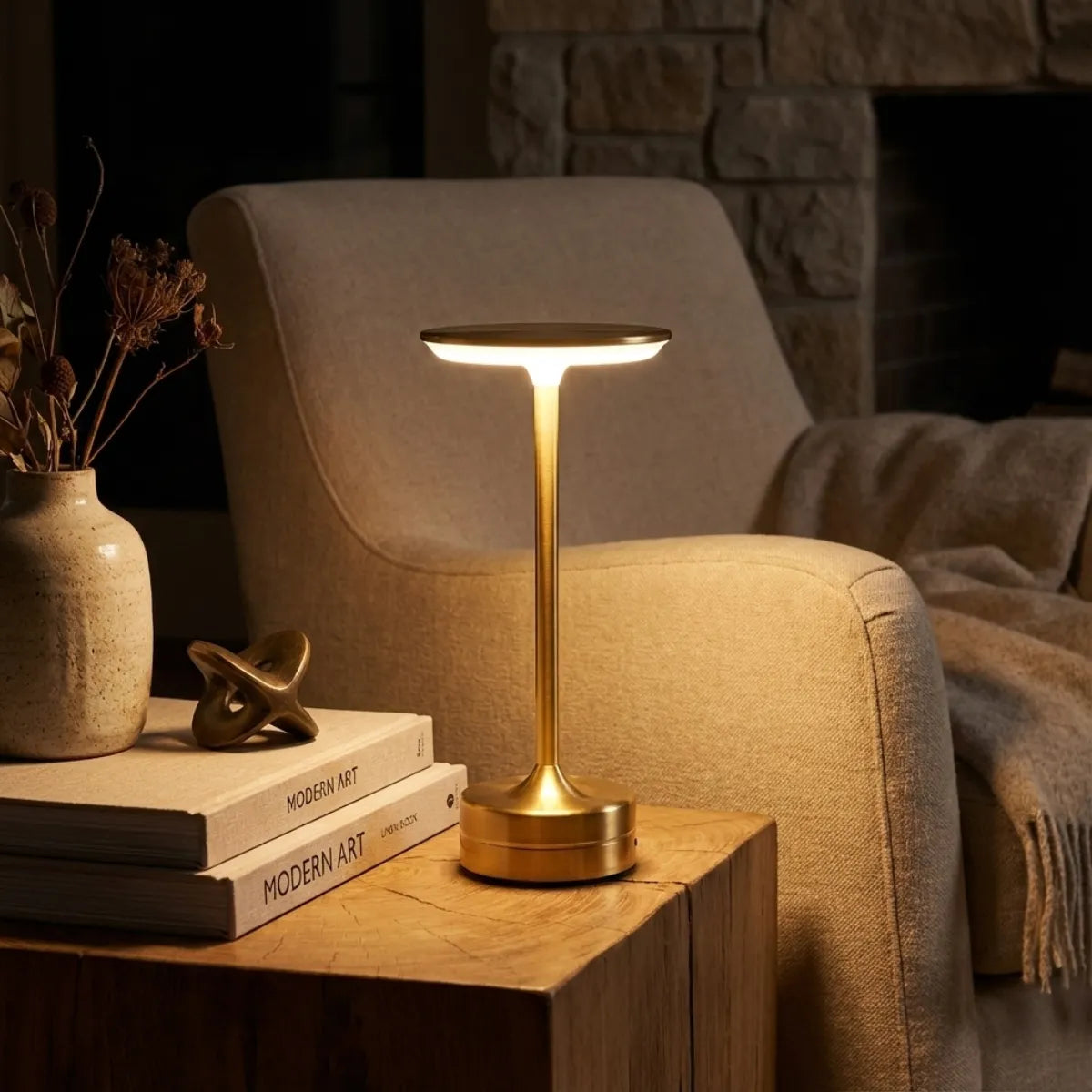 Cantinho de leitura aconchegante com luminária Majestic Dourada sobre mesa lateral de madeira maciça, ao lado de poltrona bege confortável.