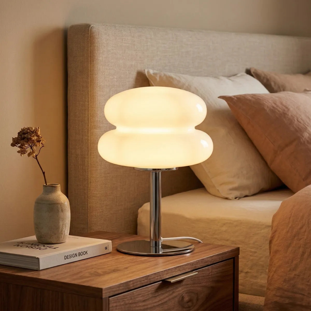 Luminária de mesa Orbe iluminando suavemente uma mesa de cabeceira de madeira. Ao fundo, uma cama com enxoval de linho em tons de bege e terracota, criando um clima de quarto aconchegante.