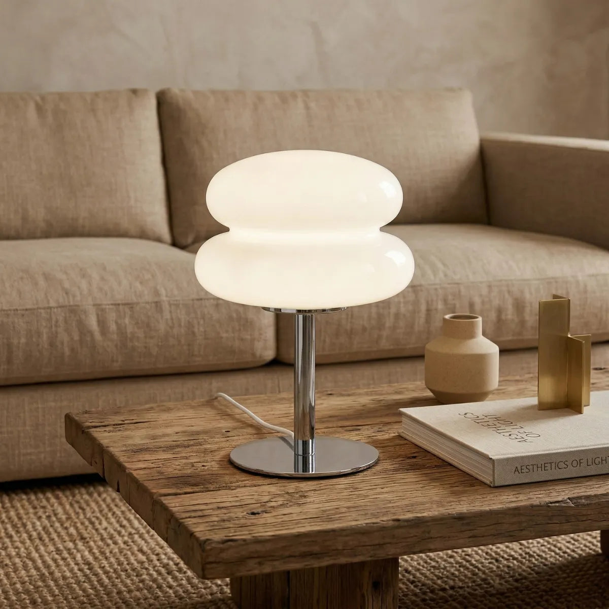 Luminária Orbe acesa sobre uma mesa de centro de madeira rústica, ladeada por um livro de estética e objetos decorativos dourados, em uma sala de estar elegante com sofá texturizado ao fundo.