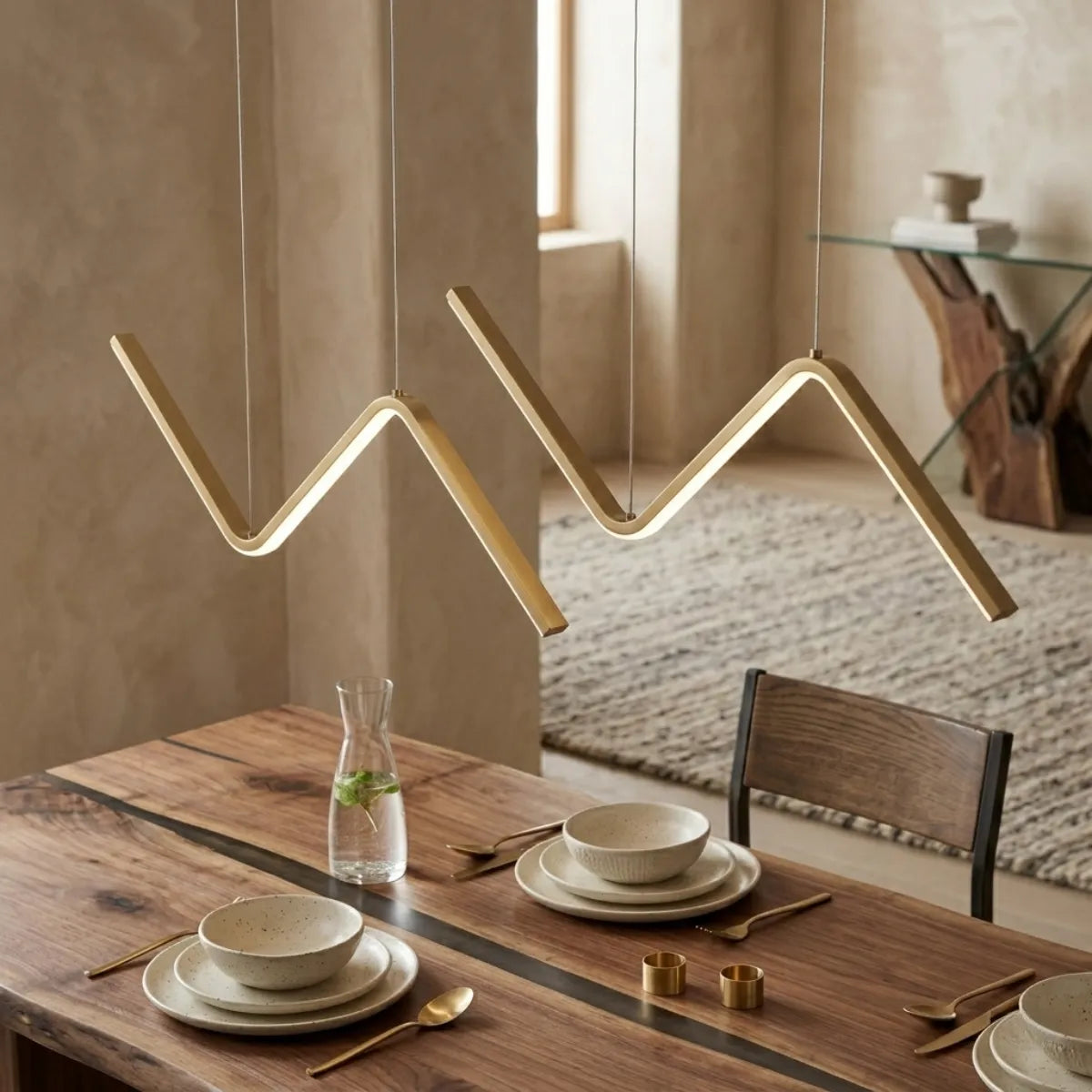 Duas luminárias pendentes douradas em zigue-zague suspensas sobre uma mesa de jantar de madeira rústica. A mesa está posta com pratos de cerâmica clara, copos e talheres dourados. O ambiente possui decoração aconchegante em tons terrosos.