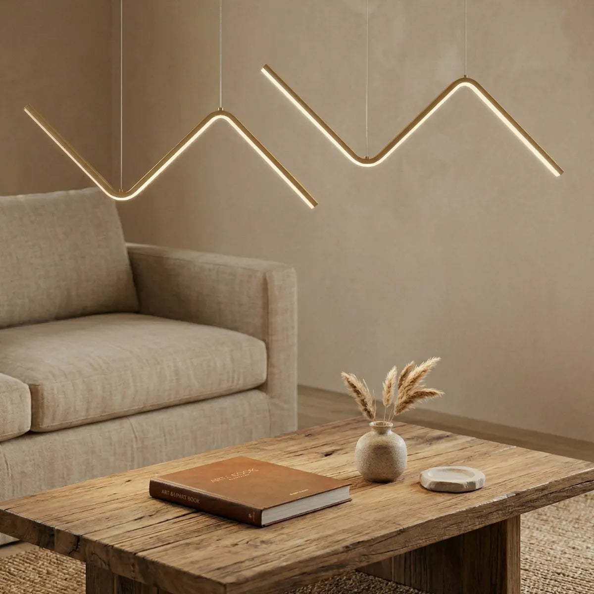 Duas luminárias pendentes douradas de design em zigue-zague ambientando uma sala de estar. Elas pairam sobre uma mesa de centro rústica de madeira crua, decorada com um livro e pequenos vasos de cerâmica. Ao fundo, um sofá bege claro.